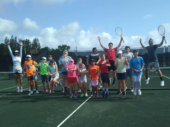 A group of kids jumping up on the tennis court in tennis camp.